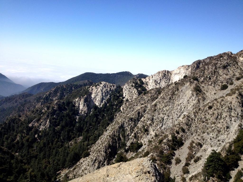 A panoramic view of rugged mountains under a clear blue sky, featuring steep rocky slopes and patches of green forestry. The lighting highlights the textures of the rock formations, with layers and cresting ridges visible in the landscape. In the distance, additional mountain ranges fade into a soft haze, creating a peaceful, natural setting. Mount Lowe Railway mountain bike trail.