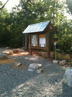 A wooden information board with a metal roof, displaying various notices and maps, situated in a natural setting with trees in the background. The ground is covered with gravel and there are a few stones positioned nearby. Coldwater Mountain mountain bike trail.