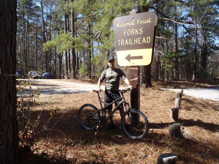 A person standing next to a "Forks Trailhead" sign in a national forest, holding a mountain bike. The background features tall trees and a gravel path, with some vehicles visible in the distance. The setting is sunny, suggesting a pleasant outdoor environment. Forks Area Trail System (FATS) mountain bike trail.