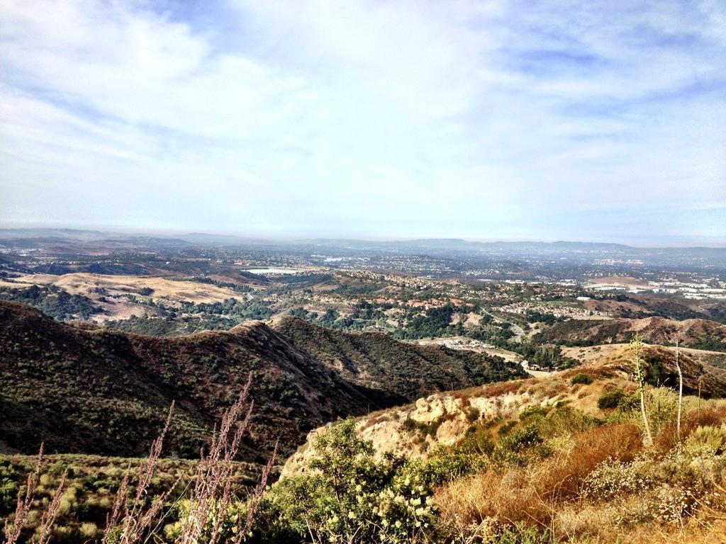 A panoramic view of rolling hills and valleys under a cloudy sky, showcasing a mix of greenery and dry vegetation, with a distant cityscape visible in the background. The Luge mountain bike trail.