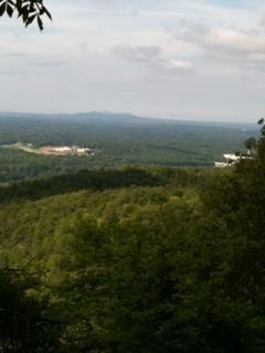 A panoramic view of a lush green landscape with rolling hills extending into the distance under a cloudy sky. The foreground features dense trees, while a low-lying area with structures can be seen in the middle ground, leading to more distant mountains. Coldwater Mountain mountain bike trail.