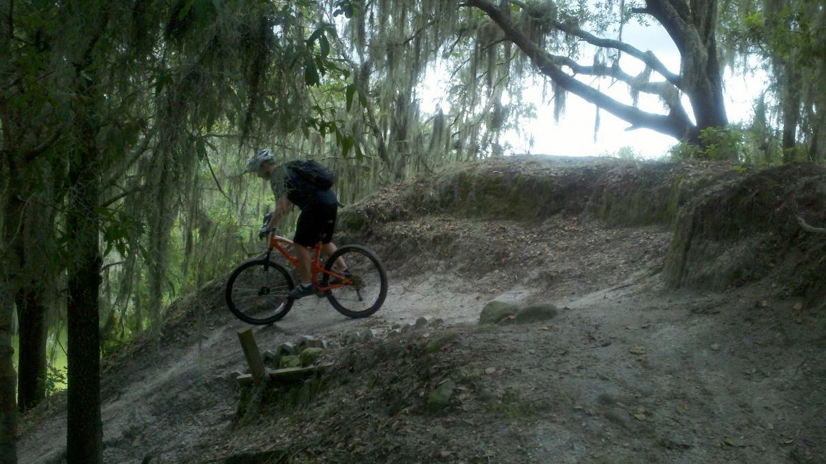 A mountain biker riding along a dirt trail surrounded by trees covered in Spanish moss. The biker is navigating a slight incline, with a backpack on their back and wearing a helmet. The scene is set in a lush, green environment typical of wooded trails. Alafia River State Park mountain bike trail.