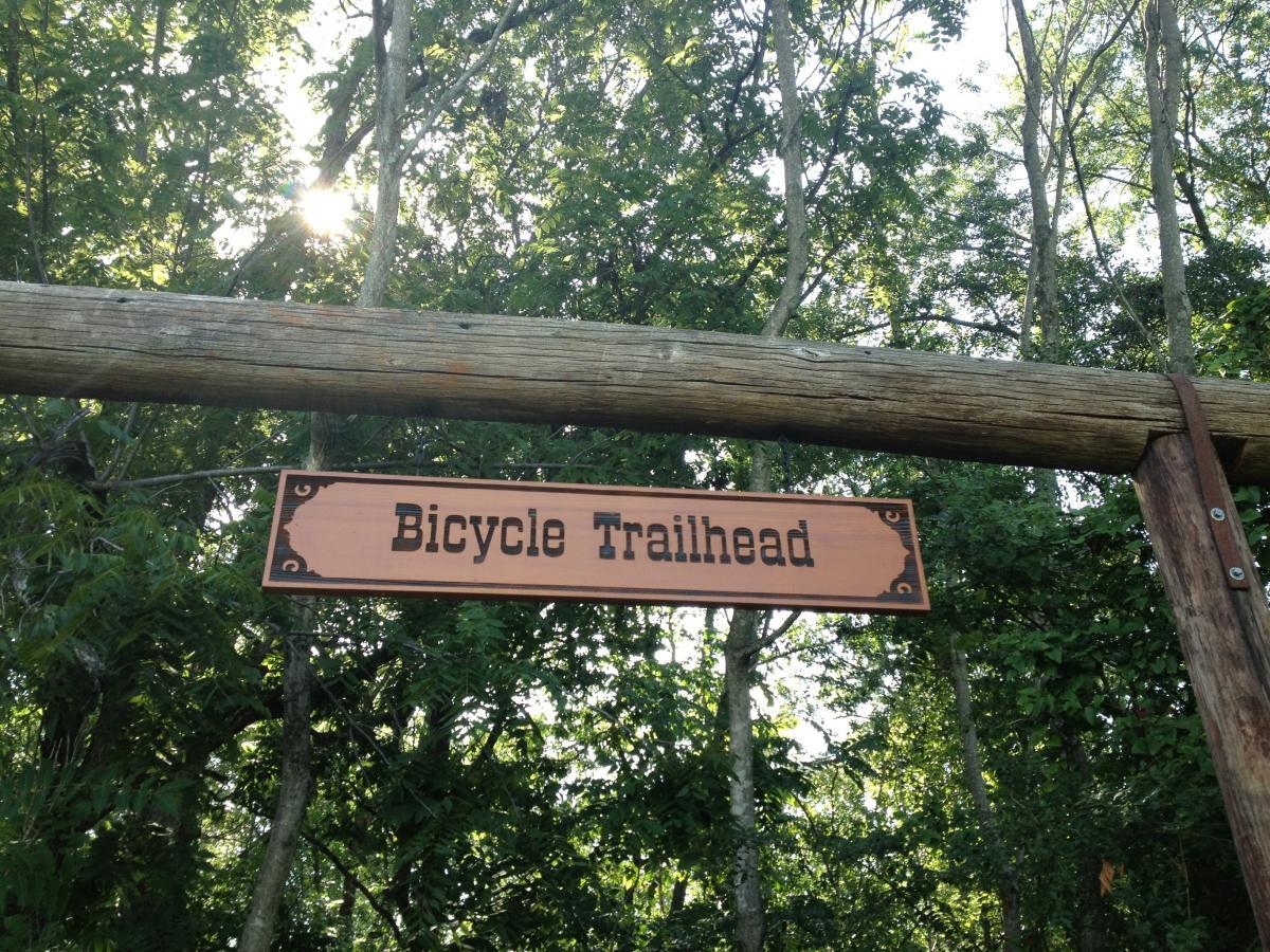 Sign for a bicycle trailhead, made of wood, hanging between two posts in a lush green forest. The sign features the text "Bicycle Trailhead" in bold letters, with sunlight filtering through the trees in the background. Heritage Park mountain bike trail.