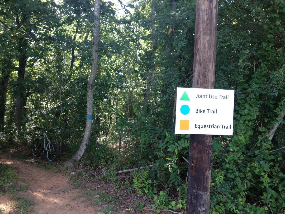 Alt text: A sign at a trailhead in a wooded area, indicating different types of trails. The sign features symbols for a Joint Use Trail (triangle), Bike Trail (circle), and Equestrian Trail (square) on a wooden post. A bicycle is leaned against a nearby tree. Heritage Park mountain bike trail.