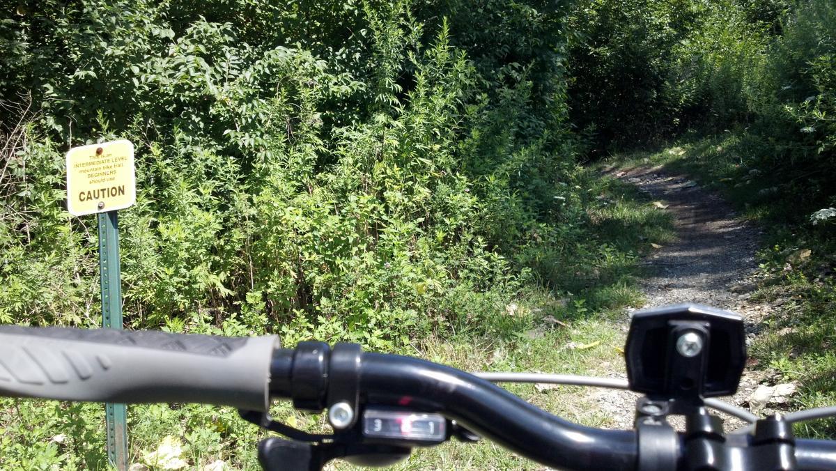 A view from a bicycle handlebar showing a narrow, wooded trail ahead, with a caution sign indicating that the trail is an intermediate-level mountain biking path. Lush greenery surrounds the path, suggesting a natural outdoor setting. Ohio And Erie Canal mountain bike trail.