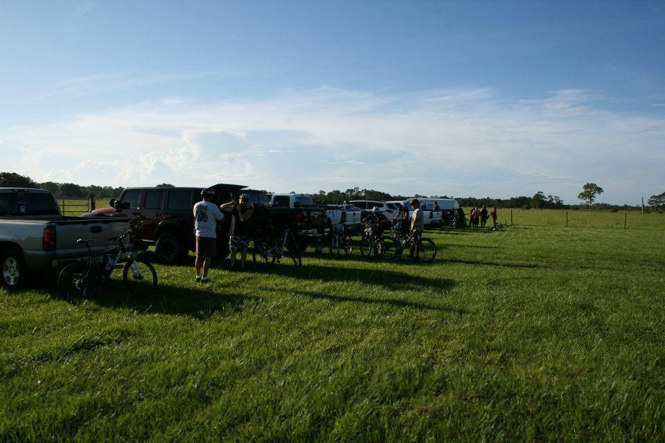 A line of parked vehicles, including trucks and SUVs, set in a grassy field under a clear blue sky. Several people are gathered near the vehicles, some with bicycles, suggesting a recreational outing. Grassy Island Trail mountain bike trail.