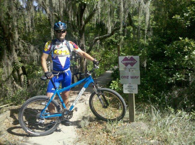 A mountain biker in a colorful cycling jersey and helmet stands next to a trail sign in a forested area. The sign indicates a one-way, very difficult trail. The biker is positioned next to a blue bike on a sandy path surrounded by greenery and hanging moss. Alafia River State Park mountain bike trail.