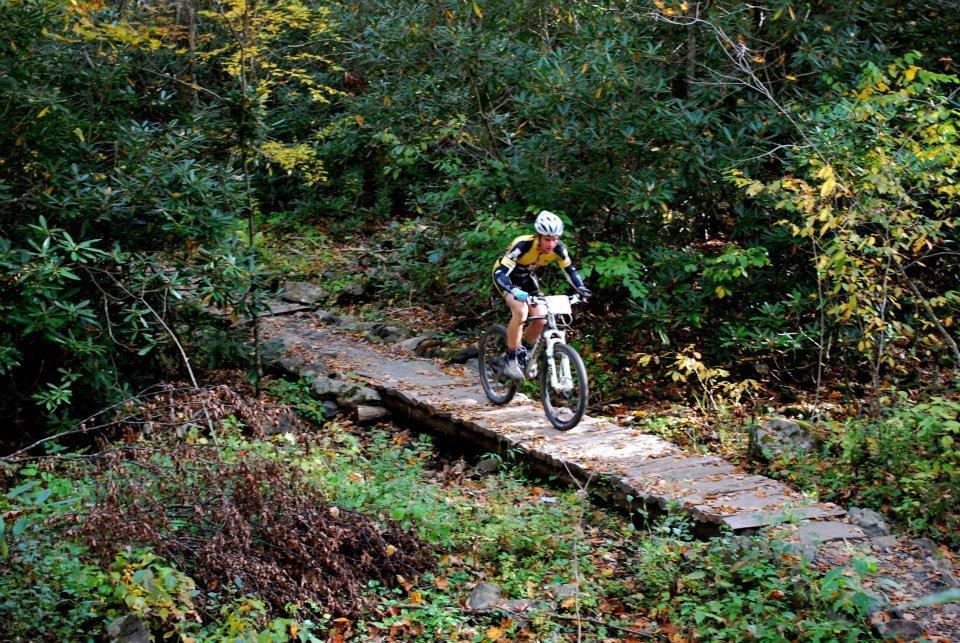 A cyclist riding a mountain bike on a stone pathway through a forest, surrounded by lush green foliage and autumn leaves. Rocky Knob Park mountain bike trail.