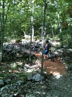 A person stands beside a mountain bike on a narrow trail surrounded by lush green trees and rocky terrain, indicating a natural outdoor setting suitable for biking. Coldwater Mountain mountain bike trail.