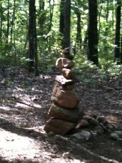 A stacked arrangement of stones in a forested area, surrounded by trees and dappled sunlight filtering through the leaves. Coldwater Mountain mountain bike trail.