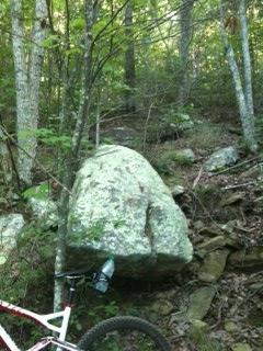 A large boulder in a dense forest, surrounded by trees and smaller rocks, with a mountain bike partially visible in the foreground. Five Points mountain bike trail.