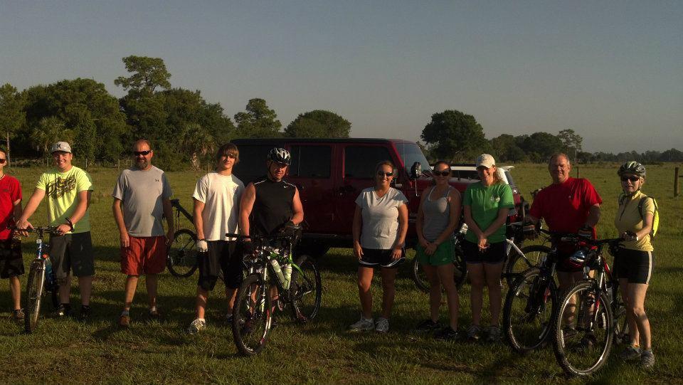 A group of eight people stands on a grassy field, posed with their bicycles beside a red vehicle. The individuals wear various athletic clothing, including shorts and t-shirts, and some have helmets. The background features trees and a clear sky, indicating a sunny day. Grassy Island Trail mountain bike trail.
