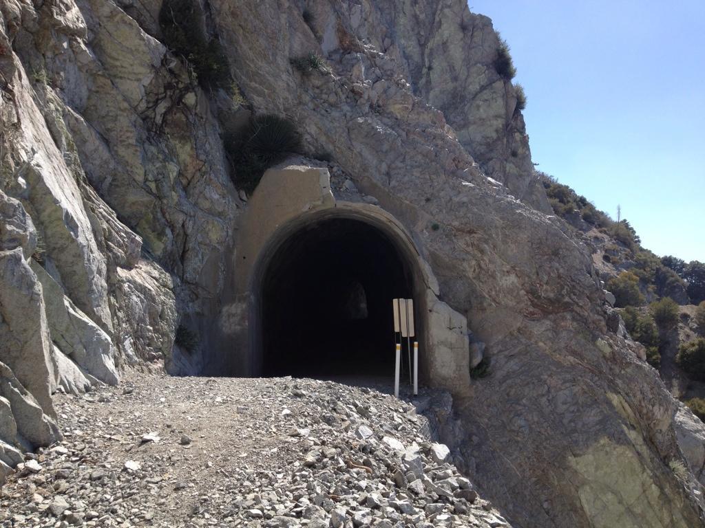 A rocky tunnel entrance on a mountainside, leading into a dark void. The tunnel is framed by rough, natural stone, with a clear path made of gravel leading up to it. Two vertical markers with black and yellow stripes are positioned next to the entrance, indicating caution. Bright blue sky can be seen in the background, highlighting the rocky terrain. Mount Lowe Railway mountain bike trail.
