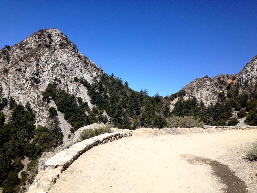 A scenic view of rocky mountains under a clear blue sky, with a dirt path curving along the foreground bordered by a stone wall. Dense greenery is visible on the slopes of the mountains, showcasing a mix of trees and shrubs. Mount Lowe Railway mountain bike trail.