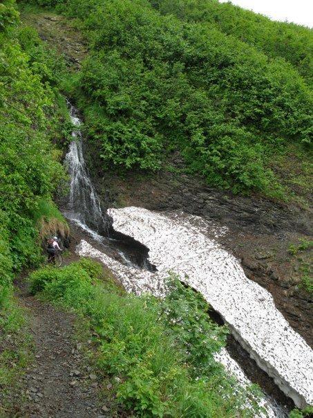 A serene landscape featuring a small waterfall cascading down a rocky slope, surrounded by lush green vegetation. The water flows over a patch of snow or ice, creating a striking contrast with the vibrant greenery. A person can be seen sitting on the ground near the waterfall, enjoying the tranquil natural setting. Crescent Lake mountain bike trail.