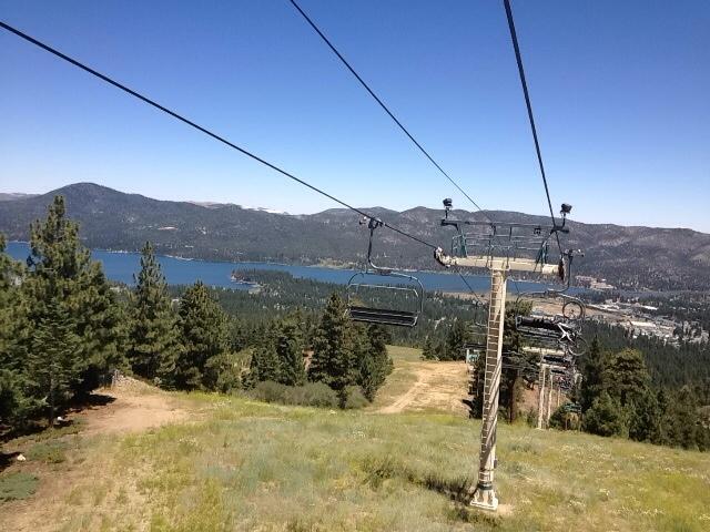 View from a ski lift overlooking a scenic landscape with a lake and mountains in the background, surrounded by trees and a clear blue sky. Big Bear Mountain Resort mountain bike trail.