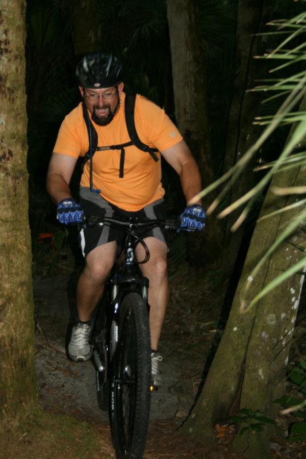 A man in an orange shirt and black shorts mountain biking through a narrow, wooded trail. He is wearing a helmet and blue gloves, showing an expression of excitement as he navigates the path surrounded by tall trees and greenery. Grassy Island Trail mountain bike trail.