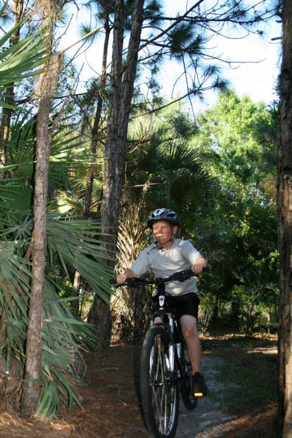 A child riding a mountain bike on a dirt path surrounded by trees and greenery. The child is wearing a helmet and casual clothing, showcasing an outdoor activity in a natural setting. Grassy Island Trail mountain bike trail.