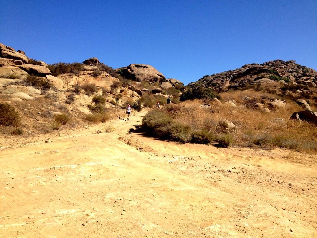 A rocky trail leading up a hillside with sparse vegetation and dry grass under a clear blue sky. Two hikers can be seen ascending the path, with rugged terrain and large boulders in the background. Hummingbird mountain bike trail.