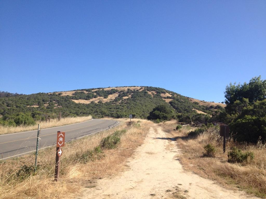 A dirt path leading towards a hillside, with a paved road on the left. The scene features dry grass and shrubbery on either side of the path, under a clear blue sky. A trail marker indicating a bike route is visible on the left. Anthony Chabot Regional Park mountain bike trail.