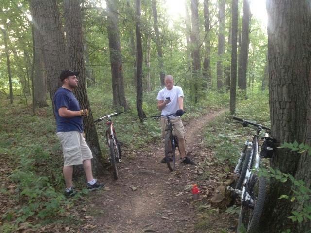 Two cyclists stand on a dirt path in a wooded area. One man, wearing a blue shirt and gloves, leans against a tree while the other, dressed in a white shirt and shorts, checks his phone. Their bikes are parked nearby on either side of the path, surrounded by lush greenery and tall trees. 3rd Battle Of Winchester Trail mountain bike trail.
