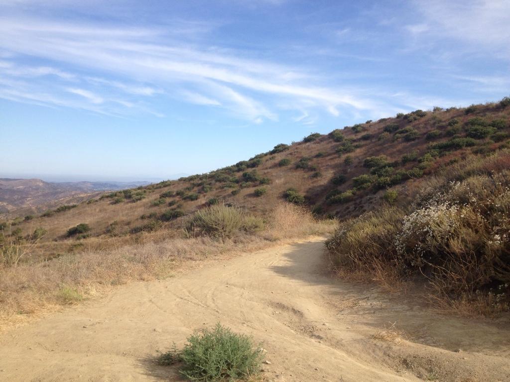 A dirt path winding through a hilly landscape with dry grass and sparse vegetation. The hills are gently sloping and dotted with low shrubs, under a clear blue sky with wispy clouds. The view extends into the distance, revealing more hills and valleys. The Luge mountain bike trail.