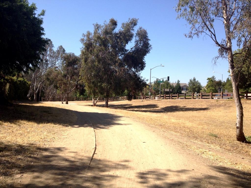 A dirt path lined with trees, leading through a dry landscape on a sunny day. In the background, a street is visible with traffic lights and a wooden fence. Fullerton Loop mountain bike trail.