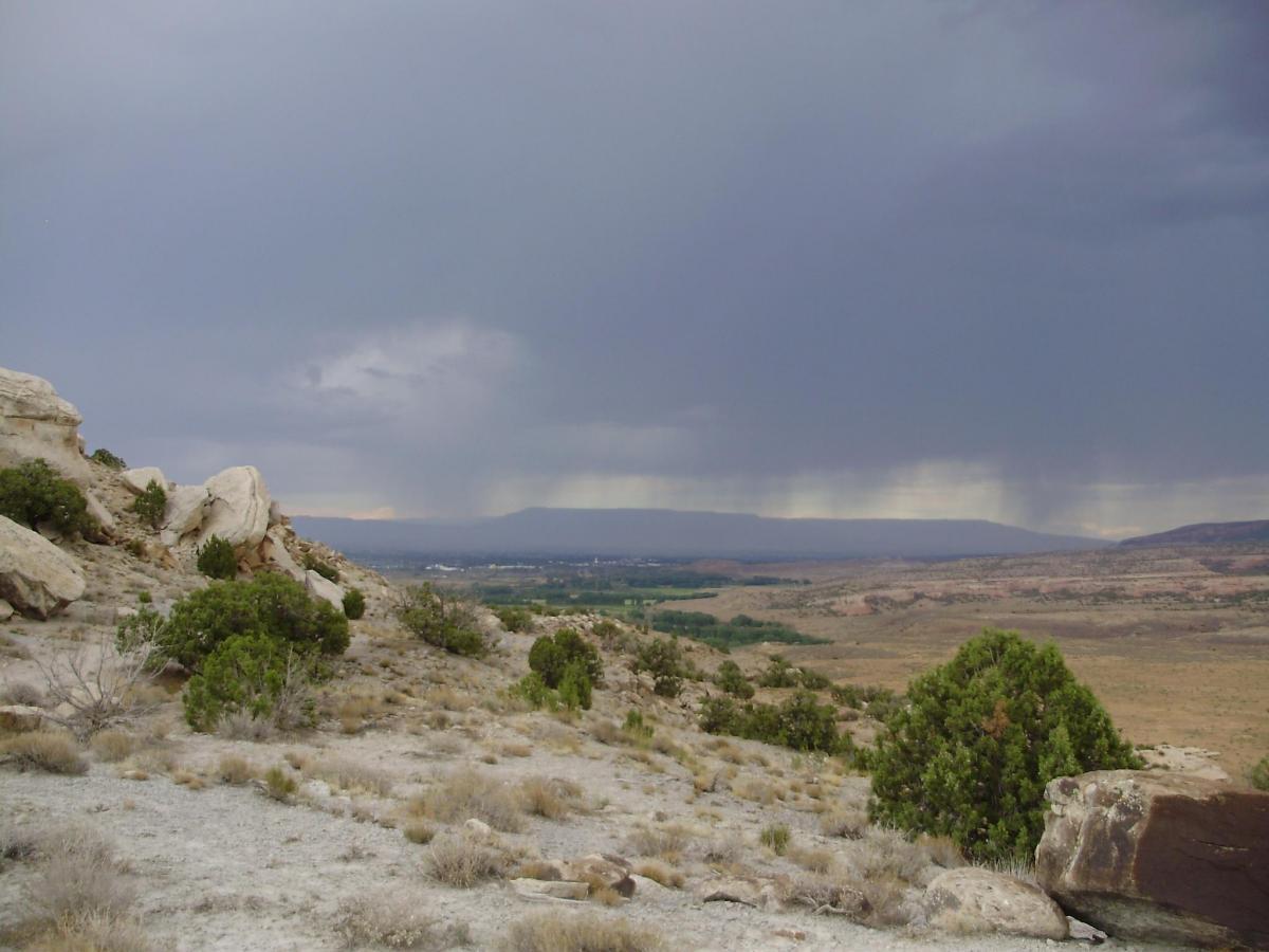 A scenic landscape featuring rolling hills and rocky outcrops under a dramatic sky filled with dark clouds. The view extends across a distant valley, showcasing patches of greenery amidst the arid terrain, hinting at the anticipation of rain in the distance. Wrangler mountain bike trail.