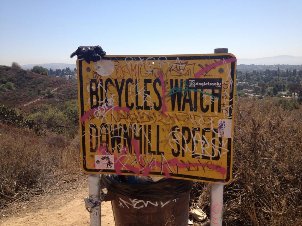A weathered yellow sign reading "Bicycles Watch Downhill Speed" with various graffiti markings, placed near a trash can on a dirt path surrounded by dry vegetation and distant hills. The background features a blue sky and scattered trees. Fullerton Loop mountain bike trail.
