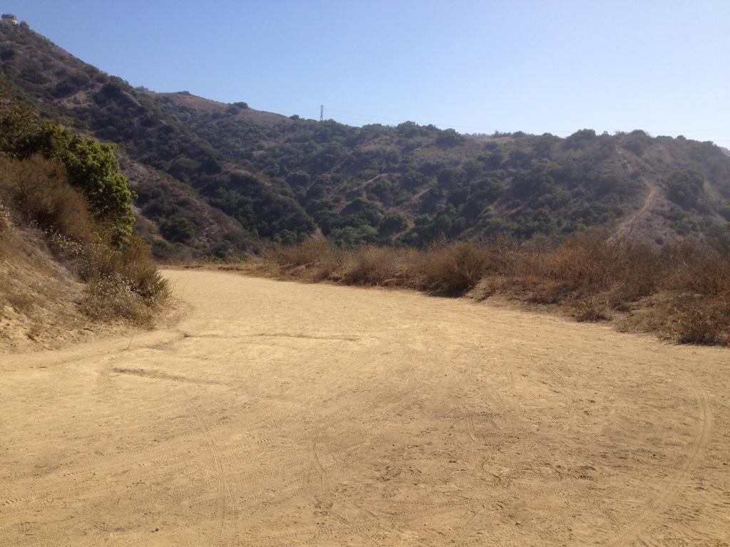A sandy dirt path winding through hilly terrain covered with dry brush and scattered greenery, under a clear blue sky. Sumac mountain bike trail.
