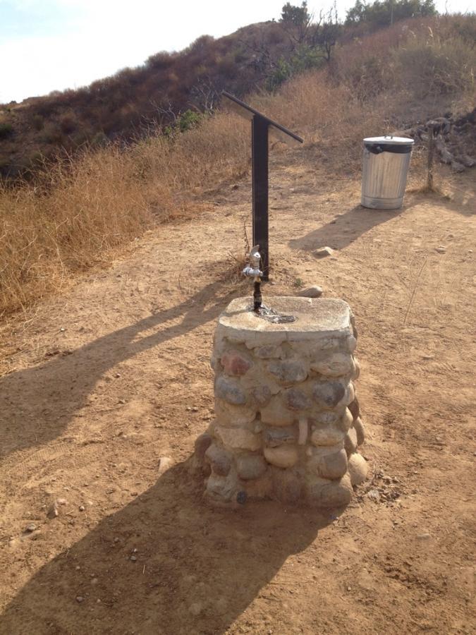 A rustic stone water fountain with a metal handle, situated on a dirt path. The surrounding landscape features dry grass and shrubs, with a trash can located nearby. The scene is illuminated by natural sunlight, creating shadows on the ground. Water Tank Rd mountain bike trail.