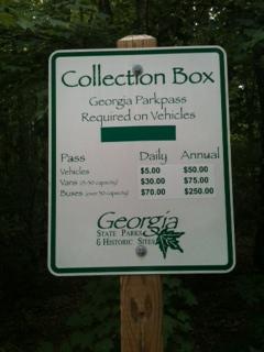 Sign for a collection box detailing Georgia Parkpass fees for vehicles and daily/annual passes. The sign indicates daily passes cost $5.00, annual passes $50.00, and other pricing for specific permits, set against a wooded background. Five Points mountain bike trail.
