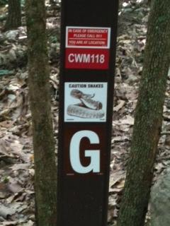 A wooden post in a forested area displaying multiple signs. The top sign reads "In case of emergency please call 911. You are at location CWM118." Below that is a warning sign stating "Caution Snakes," featuring an illustration of a snake. At the bottom of the post, there is a large letter "G." The surroundings are wooded with fallen leaves on the ground. Coldwater Mountain mountain bike trail.