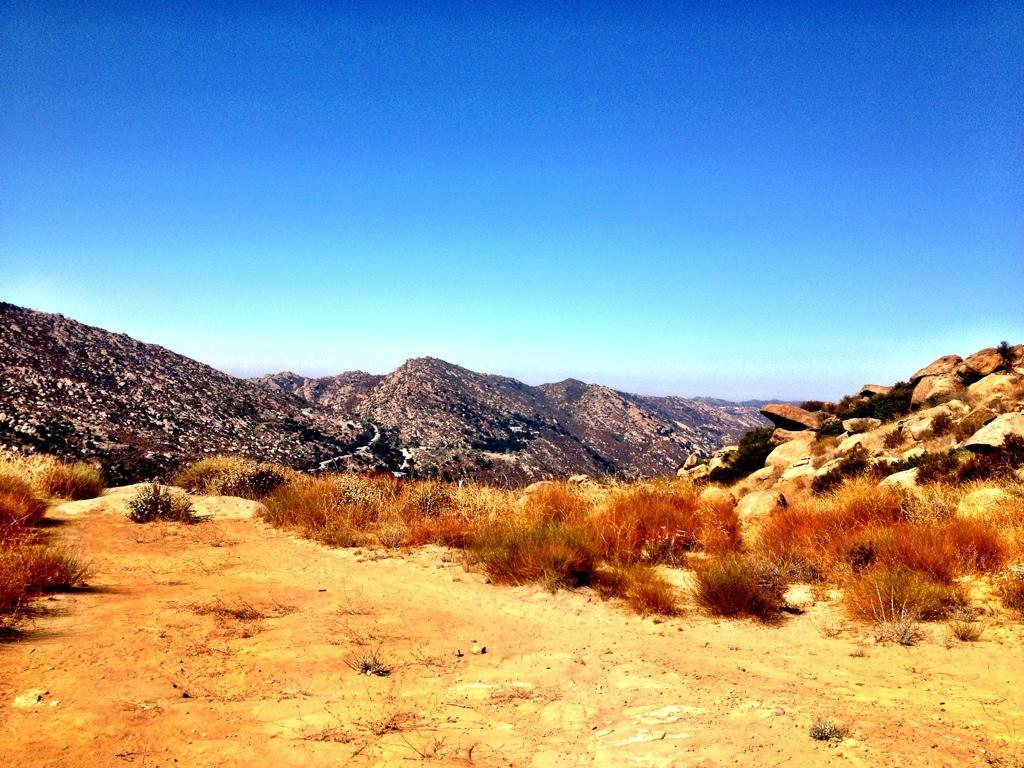 A panoramic view of mountains under a clear blue sky, featuring sandy terrain and patches of dry grass. The landscape showcases rolling hills and rocky formations, creating a serene natural setting. Hummingbird mountain bike trail.