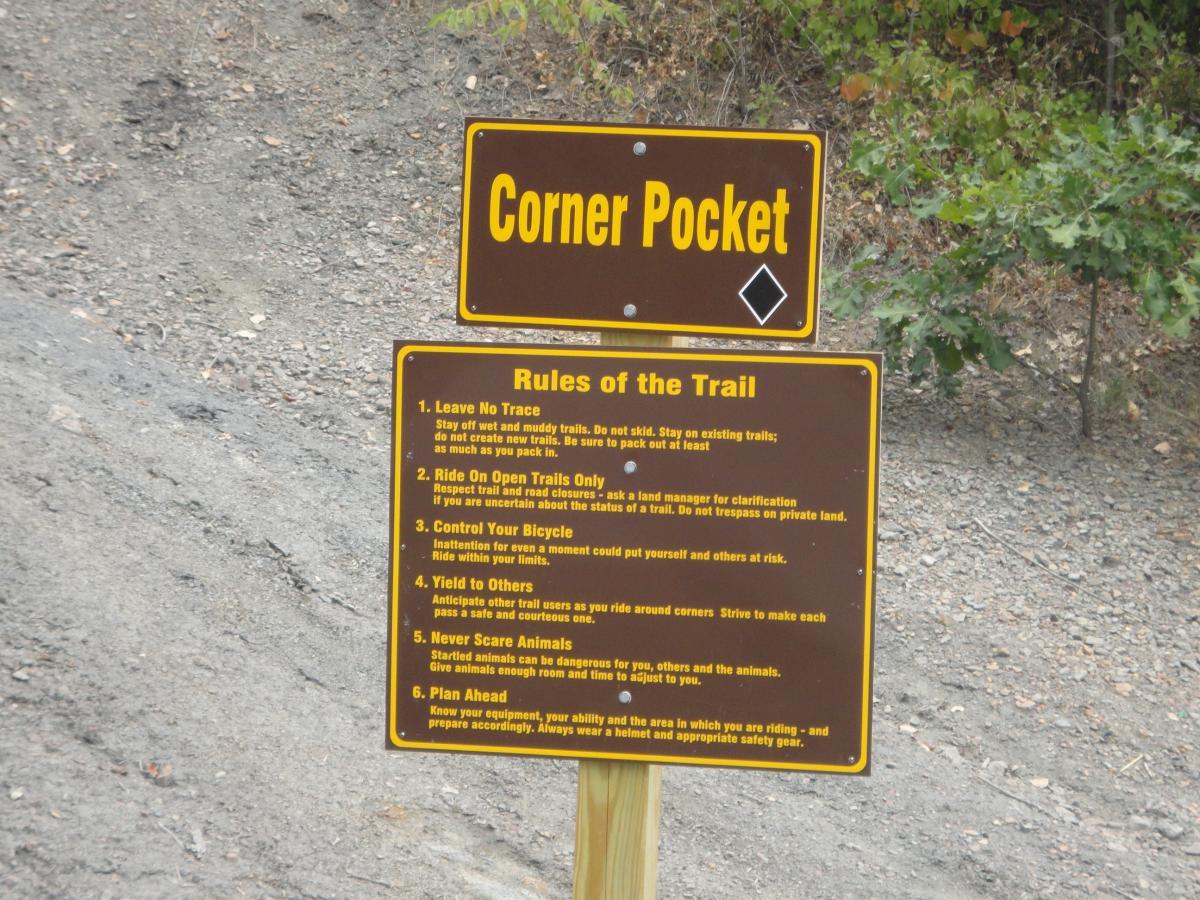 A sign titled "Corner Pocket" with a list of trail rules, including guidelines such as "Leave No Trace," "Ride On Open Trails Only," and other safety reminders for trail users. The sign is situated in a natural outdoor setting, with a dirt pathway and surrounding greenery. Banner Lakes At Summerset Park mountain bike trail.