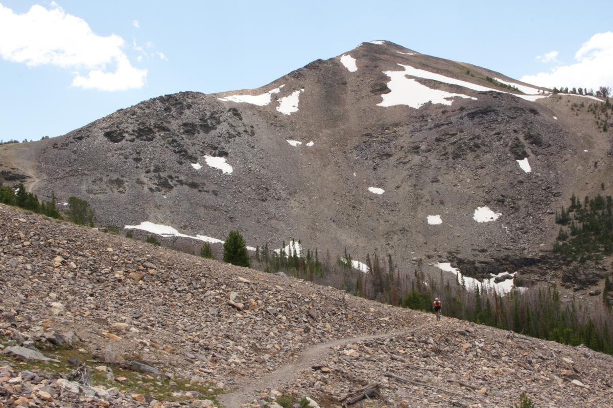 A scenic view of a rugged mountain landscape with rocky terrain, featuring patches of snow on the slopes. A lone hiker can be seen on a winding trail, surrounded by sparse trees and boulders, under a clear blue sky. Rocky Creek Loop mountain bike trail.