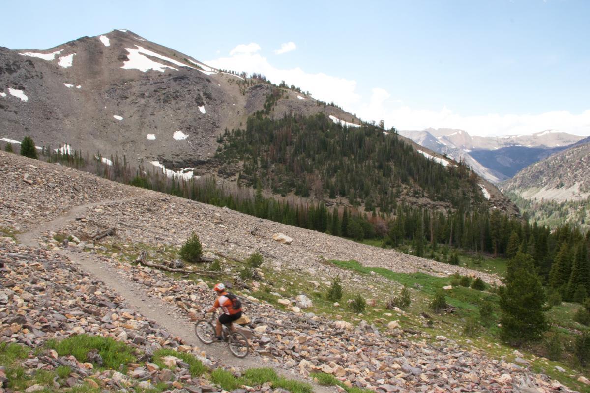 A mountain biker riding along a rocky trail in a mountainous area, surrounded by greenery and trees, with snow-capped peaks in the background under a partly cloudy sky. Rocky Creek Loop mountain bike trail.