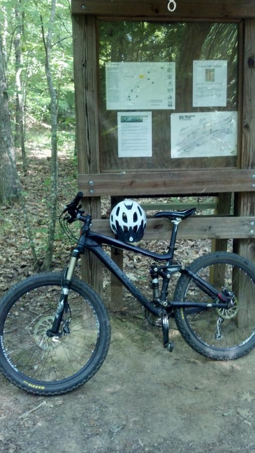 A black mountain bike and a white helmet are leaning against a wooden trail information board in a wooded area. The board features maps and information about the surrounding trails, with trees and greenery visible in the background. Oak Mountain State Park Bump Trail mountain bike trail.