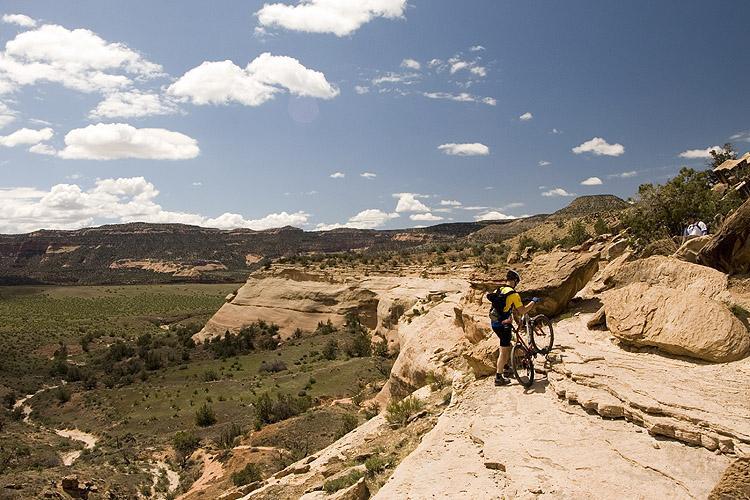 A mountain biker standing on a rocky trail, holding a bicycle, with expansive views of rolling hills and dramatic cliffs in the background under a partly cloudy sky. Colorado National Monument mountain bike trail.