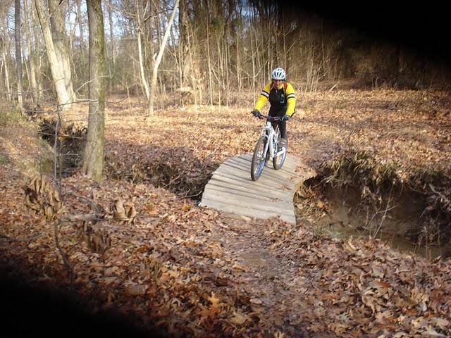 A cyclist riding a mountain bike across a wooden bridge over a small ravine in a wooded area, surrounded by fallen leaves and trees. The cyclist is wearing a yellow and black jacket and a helmet. Stanky Creek mountain bike trail.