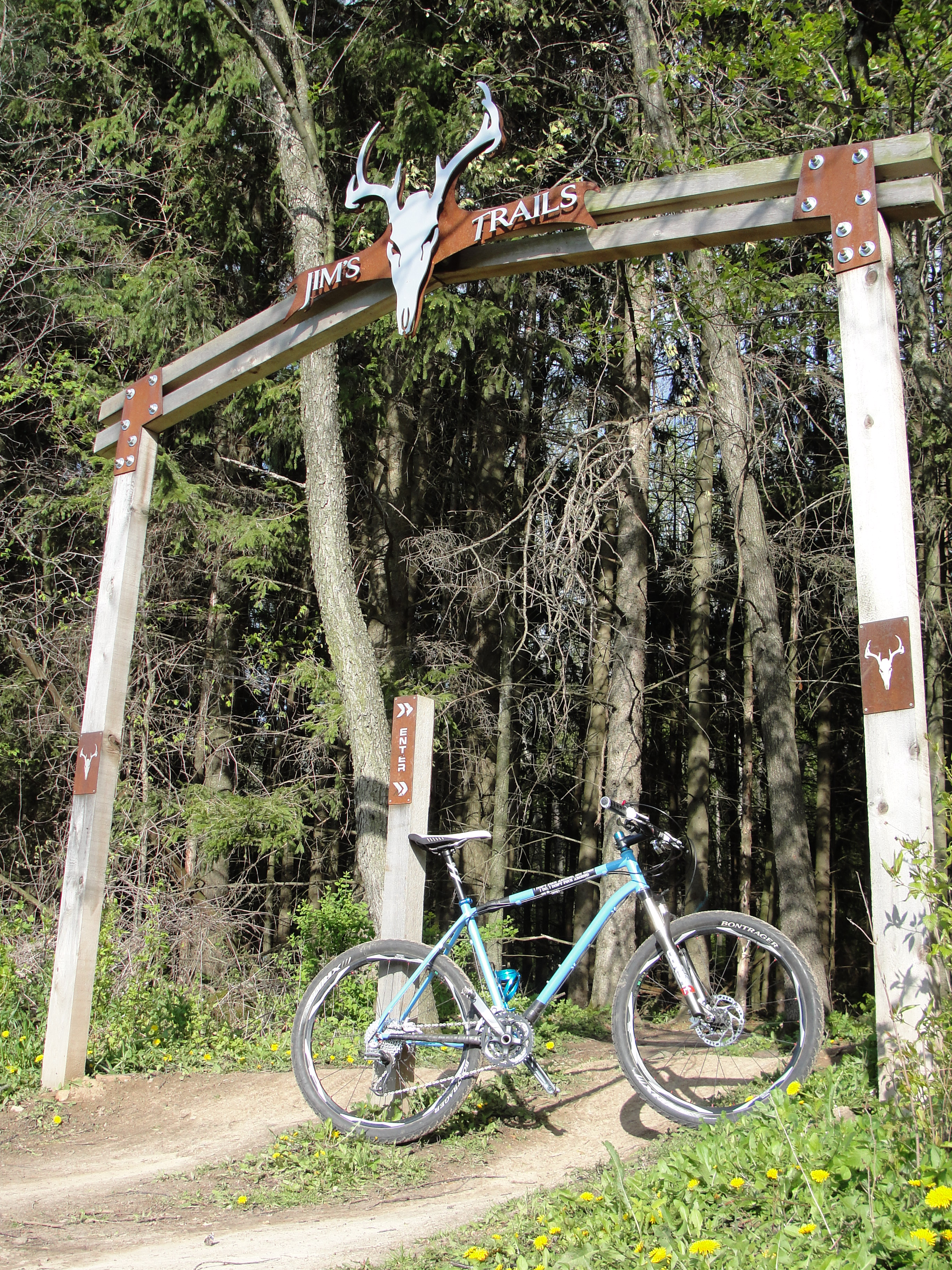 A mountain bike rests on a dirt path beneath a wooden archway labeled "Jim's Trails," adorned with a decorative skull, surrounded by lush green trees.