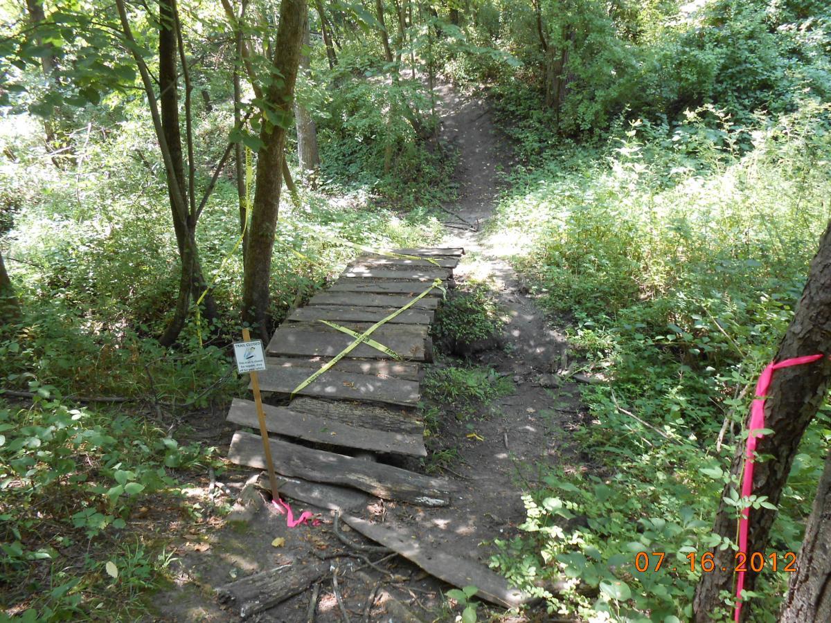 An uneven wooden bridge crossing a small gap in a wooded area, marked with yellow caution tape. A sign indicates the trail is closed, and vibrant pink flagging is visible on nearby trees. Lush greenery surrounds the path, suggesting a dense forest environment. Beverly Park mountain bike trail.