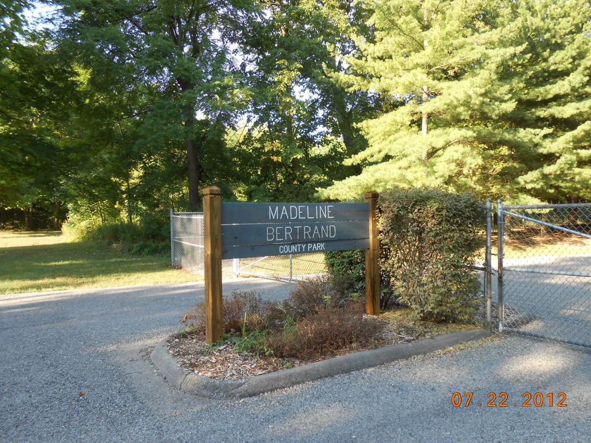 Sign for Madeline Bertrand County Park, set against a backdrop of trees, with a gravel entrance road leading into the park. The date "07.22.2012" is visible at the bottom of the image. Madeline Bertrand mountain bike trail.