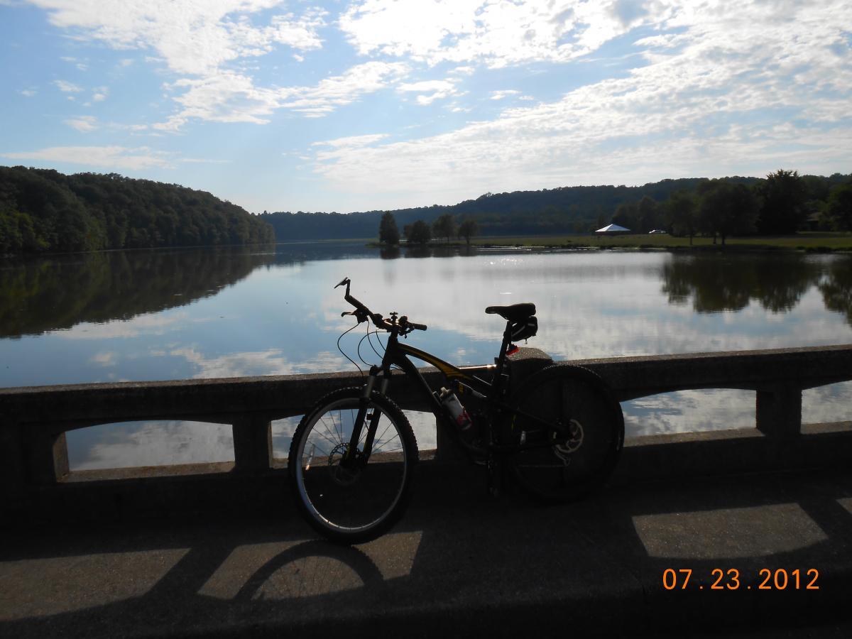 A black mountain bike resting on a stone railing, overlooking a serene lake reflecting the sky and surrounding greenery. The scene is captured on a bright day with fluffy clouds and gentle sunlight, evoking a peaceful outdoor atmosphere. Date displayed: July 23, 2012. Versailles State Park mountain bike trail.