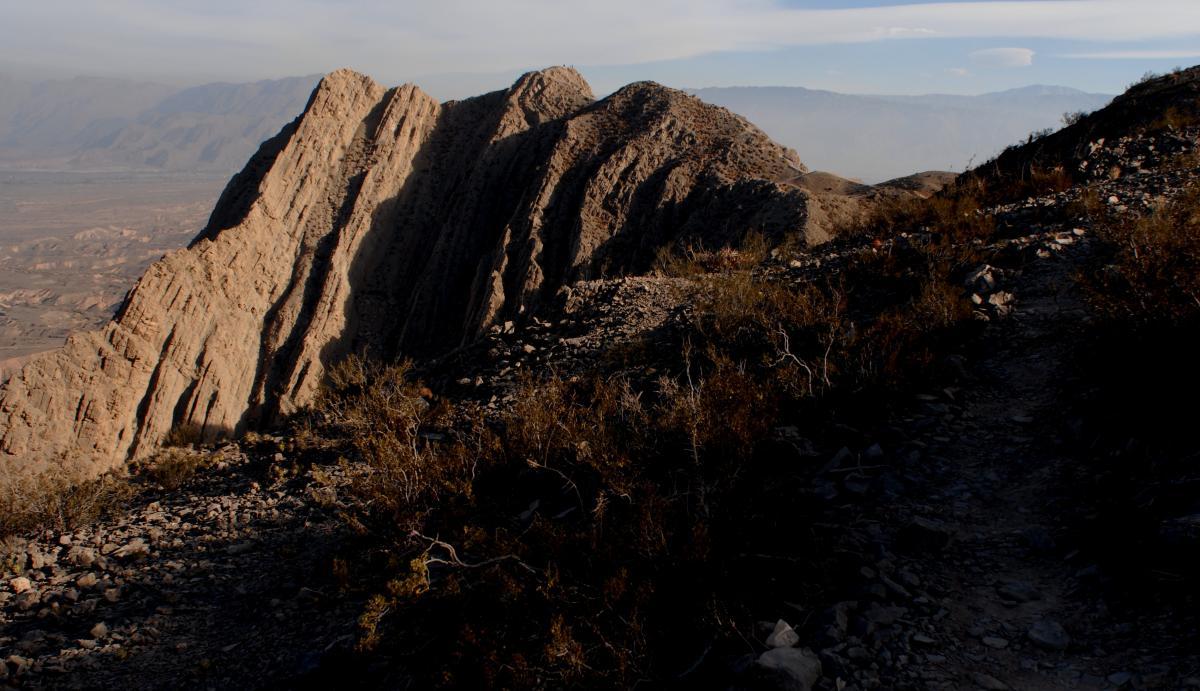 A rocky mountain landscape featuring steep, ridged cliffs illuminated by soft light, with sparse vegetation in the foreground and a distant view of rolling hills under a cloudy sky. Tres Marias mountain bike trail.