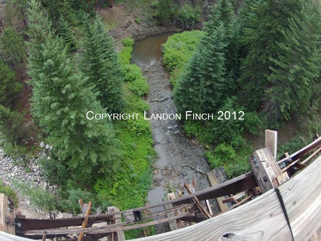 Aerial view of a winding creek surrounded by dense evergreen trees, with a wooden structure visible in the foreground. The scene captures the natural beauty of a forested area. The George S. Mickelson Trail mountain bike trail.