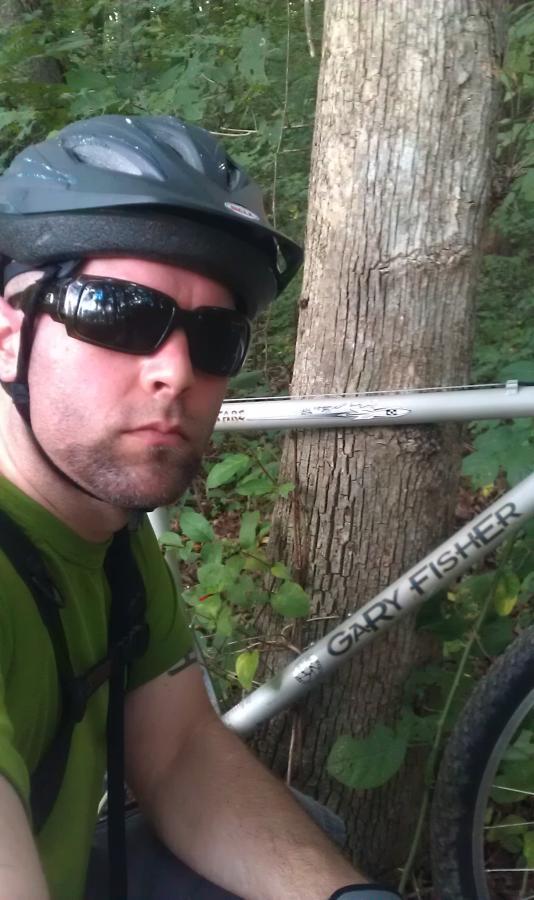 A person wearing a black helmet and sunglasses takes a selfie next to a mountain bike with a visible "Gary Fisher" label. The background features lush greenery and a tree. The individual is dressed in a green shirt and is positioned off to the side, focusing on the bike. North Meck Park mountain bike trail.