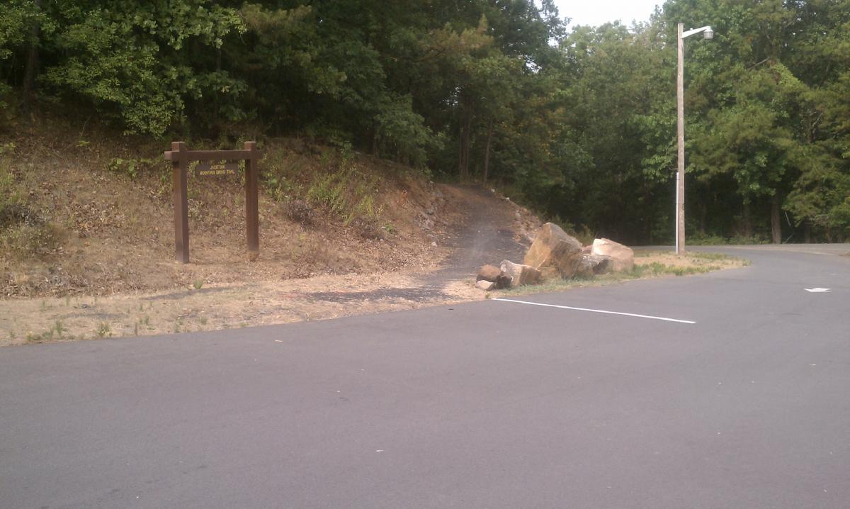 Sign for the "Mountain View Trail" near a parking area, with a dirt path leading into a wooded area. Large rocks are positioned near the path, surrounded by sparse vegetation and trees. Jackfork Trail mountain bike trail.