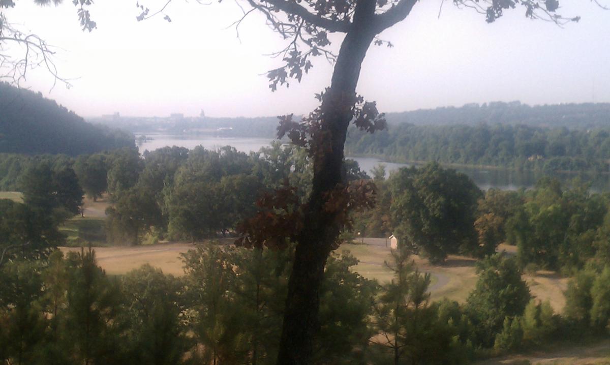 A scenic view of a river surrounded by lush greenery and hills, with a focus on a tree in the foreground. The sky is slightly hazy, suggesting an early morning or late afternoon atmosphere. Burns Park mountain bike trail.