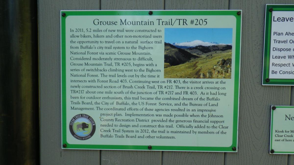 A sign describing the Grouse Mountain Trail (TR #205) in the Bighorn National Forest, detailing its 5.2 miles of trail for bikers, hikers, and non-motorized users. The sign includes information about the trail's difficulty level, route, intersections with other trails, and its development through collaboration among local agencies. An image of scenic mountains and valleys is also displayed on the sign. Clear Creek mountain bike trail.